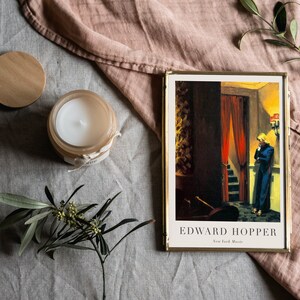May include: A framed print of a painting by Edward Hopper, titled "New York Movie". The painting depicts a woman in a blue dress standing in a doorway, looking down a staircase. The print is on a light gray surface with a wooden candle lid, a candle, and a sprig of greenery.