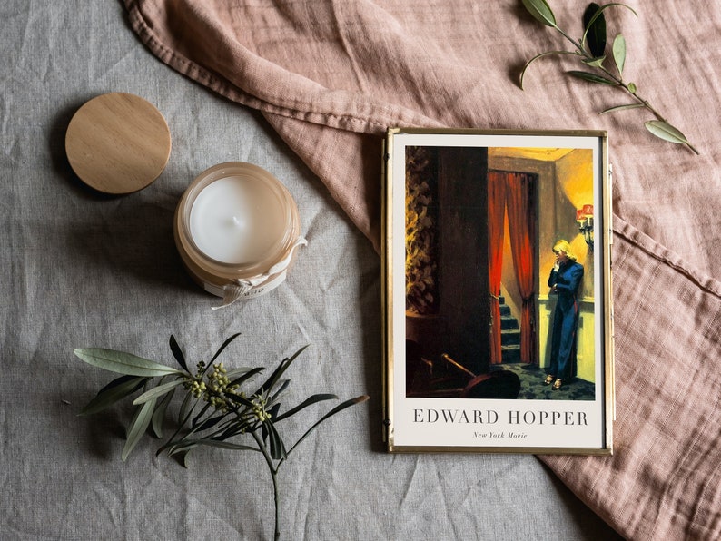May include: A framed print of a painting by Edward Hopper, titled "New York Movie". The painting depicts a woman in a blue dress standing in a doorway, looking down a staircase. The print is on a light gray surface with a wooden candle lid, a candle, and a sprig of greenery.