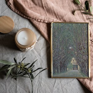 May include: A gold-framed picture with a painting of a tree-lined path leading to a building. The picture is on a light gray surface with a wooden lid, a candle, and a sprig of greenery.
