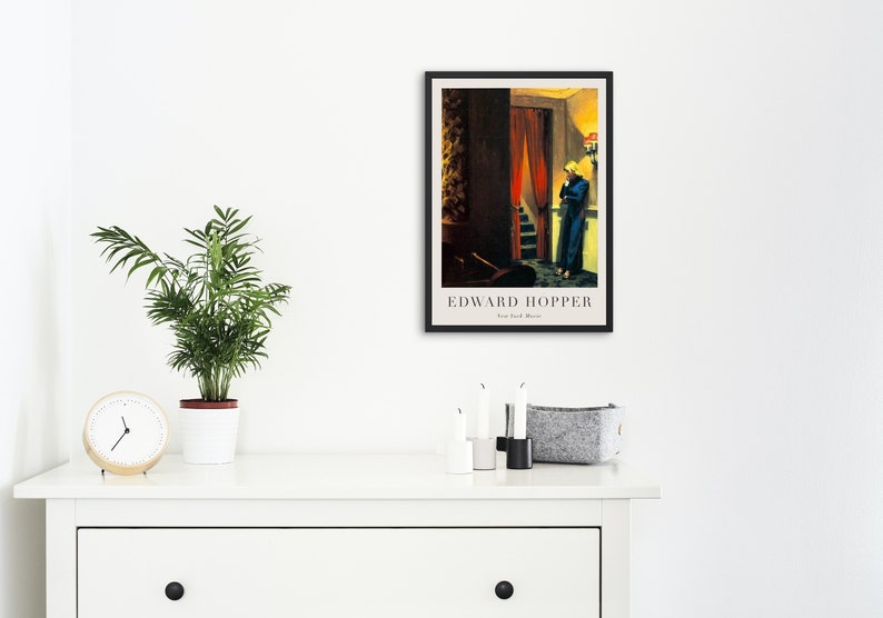 May include: A framed poster of a painting by Edward Hopper titled "New York Movie". The painting depicts a woman in a blue dress standing in a doorway with a red curtain. The woman is looking down at something in her hand.