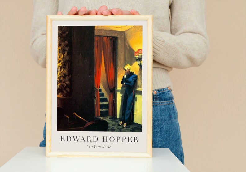 May include: A framed print of a painting by Edward Hopper titled "New York Movie". The painting depicts a woman in a blue dress standing in a dimly lit hallway with red curtains and a staircase.
