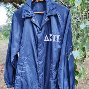 May include: A navy blue nylon jacket with a snap closure and a white embroidered Delta Sigma Pi logo on the front.