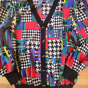 May include: A colorful vintage cardigan with a patchwork design. The button-up sweater features a black V-neck, cuffs, and hem. The pattern includes red, blue, green, and purple geometric shapes, including houndstooth and checkered patterns.