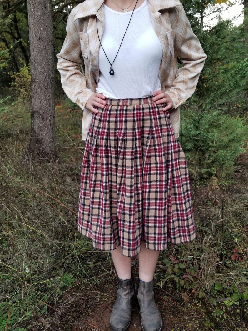 May include: A plaid skirt with a brown, red, and black pattern. The skirt is pleated and falls to the knee. The skirt is paired with a white t-shirt and a beige plaid shirt.