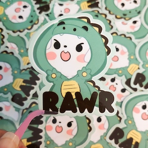 May include: A sticker featuring a cartoon dog wearing a green dinosaur costume. The dog has a white face with black eyes and a pink nose. The dinosaur costume has yellow accents and the word "RAWR" is printed in black letters below the dog.