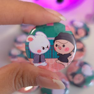 May include: A round button featuring a cartoon illustration of a white rabbit and a person in a black beanie. The background includes a green fence and a brick-paved walkway. Other buttons are visible in the background.