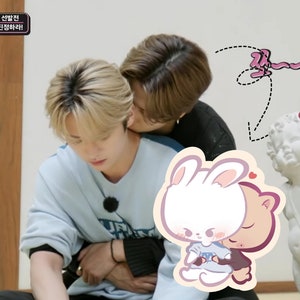 May include: Two people are hugging, one with blonde hair and a light blue sweater, the other with brown hair and a black sweater. A cartoon sticker of a white bunny hugging a brown bear is in the foreground.