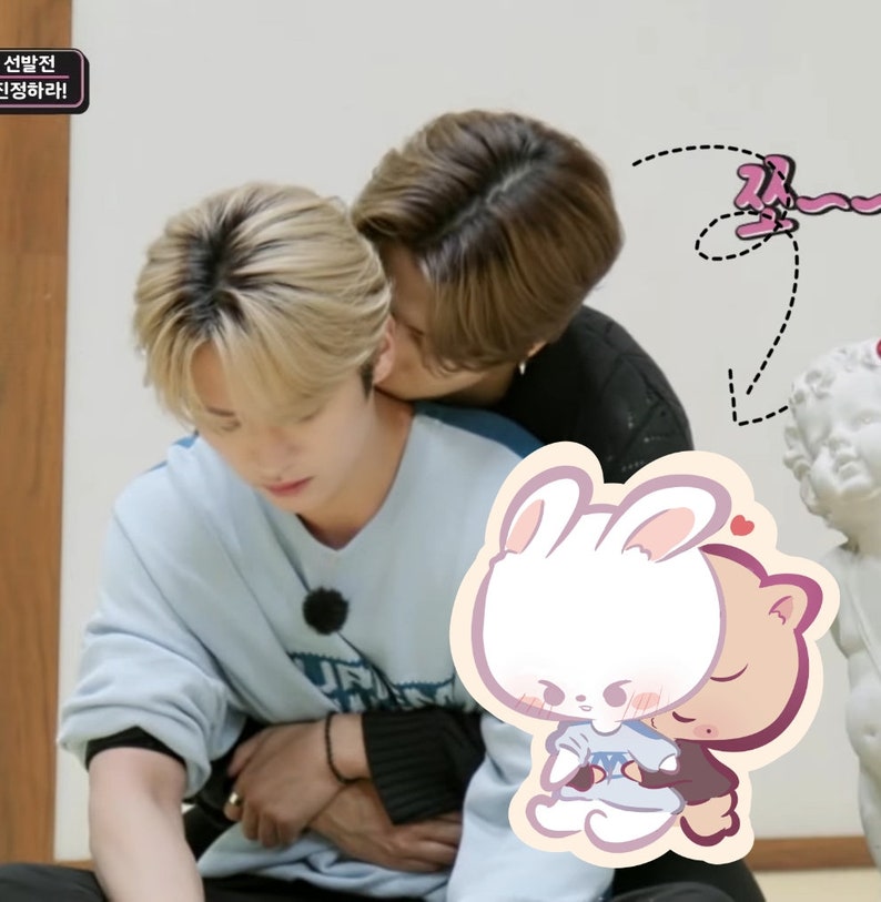 May include: Two people are hugging, one with blonde hair and a light blue sweater, the other with brown hair and a black sweater. A cartoon sticker of a white bunny hugging a brown bear is in the foreground.