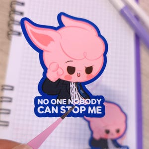 Puede incluir: Una pegatina rosa con un diseño de personaje de dibujos animados, con una cara rosa con un contorno azul y el texto "NO ONE NOBODY CAN STOP ME". La pegatina está colocada en un cuaderno de espiral.