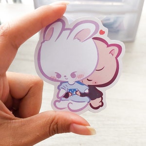 May include: A sticker featuring a white bunny with pink ears and a pink pig with closed eyes. The bunny is wearing a blue dress and the pig is wearing a black shirt. The bunny is hugging the pig and there is a small red heart above their heads.