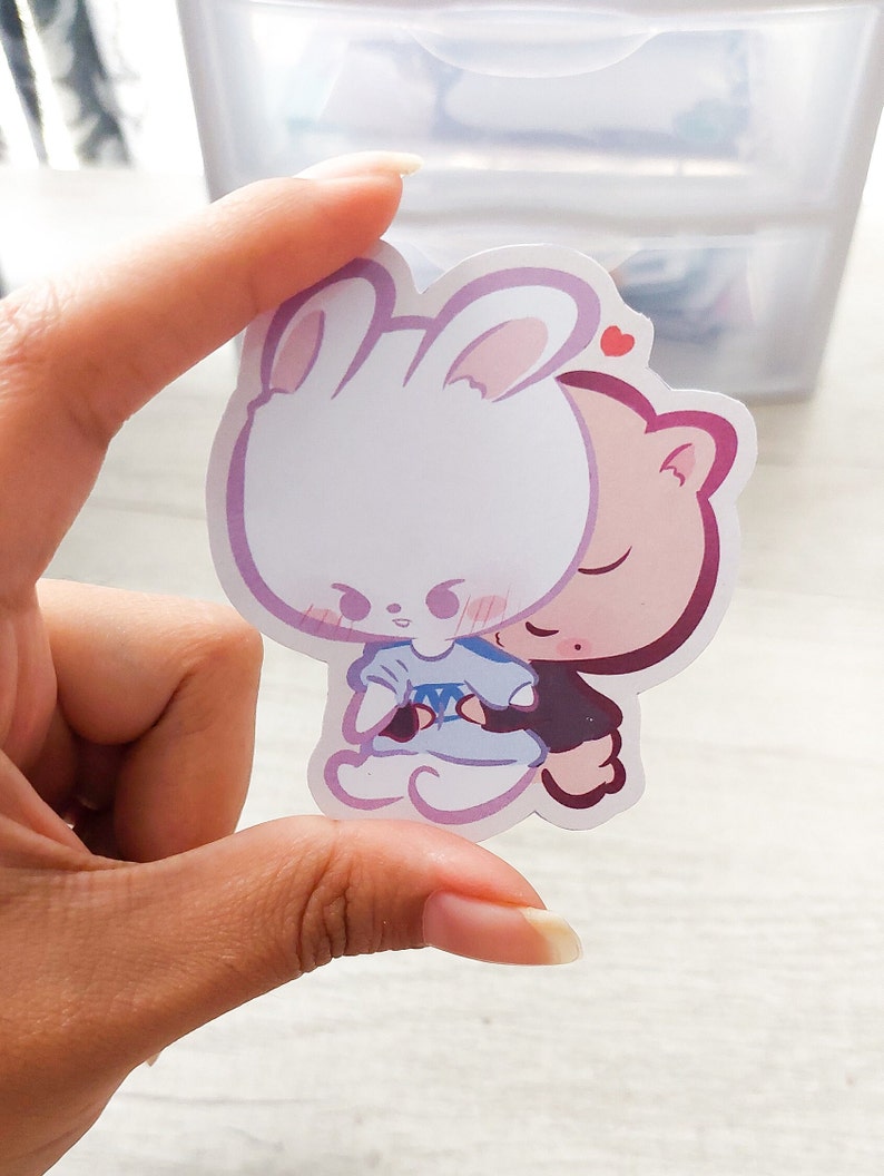 May include: A sticker featuring a white bunny with pink ears and a pink pig with closed eyes. The bunny is wearing a blue dress and the pig is wearing a black shirt. The bunny is hugging the pig and there is a small red heart above their heads.