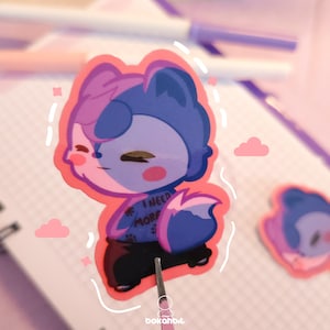 Puede incluir: Una pegatina de un animal de dibujos animados rosa y azul con un fondo rosa. El animal lleva una camisa azul con el texto "Necesito más café". La pegatina está en un cuaderno con una encuadernación en espiral.