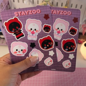 Puede incluir: Dos hojas de pegatinas con los textos "STAYZOO" y "TAYZOO" en rosa. Cada hoja presenta varias pegatinas de osos de dibujos animados con trajes rojos y rosas. Las pegatinas están sobre un fondo de cuadrícula púrpura.