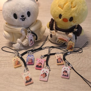Puede incluir: Una colección de pequeños y coloridos amuletos con imágenes de personas, unidos a cordones negros. Los amuletos tienen varias formas, incluyendo rectángulos y diseños de animales. Dos peluches, un oso blanco y un pollito amarillo, están en el fondo.