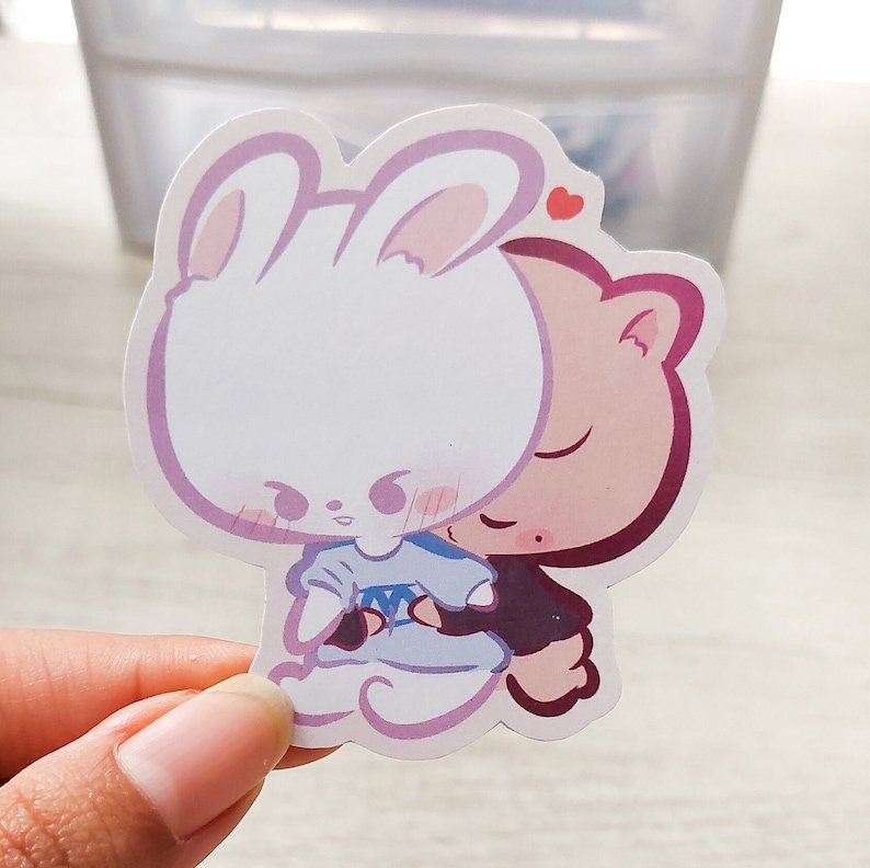 May include: A sticker featuring a white bunny with pink ears and a pink cat with closed eyes. The bunny is wearing a blue and white dress and the cat is wearing a black and white dress. The bunny is hugging the cat. The sticker has a pink heart in the top right corner.