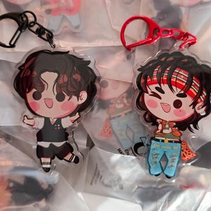 Pode incluir: Dois chaveiros de acrílico com personagens de desenhos animados. Um chaveiro tem um fecho preto e retrata um personagem com colete e calções pretos. O outro chaveiro tem um fecho vermelho e mostra um personagem com uma bandana e jeans rasgados.