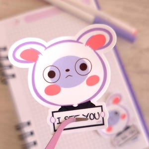 Puede incluir: Una pegatina de oso de dibujos animados blanco y morado con el texto "I SEE YOU" sujeta con unas pinzas rosas. La pegatina tiene orejas y mejillas rosas y está colocada en un cuaderno con un bolígrafo en el fondo.