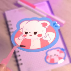 Puede incluir: Un oso de dibujos animados blanco que lleva un salvavidas a rayas rosa y blanco con un contorno azul. El oso tiene una expresión preocupada y está sudando. La pegatina está en una libreta rosa y blanca.