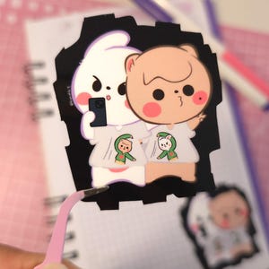 Puede incluir: Una pegatina troquelada con un conejo blanco y un oso pardo, ambos con camisetas a juego con personajes de dibujos animados. El conejo sostiene un teléfono, haciéndose un selfie. La pegatina es negra con borde blanco.