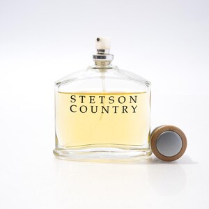 May include: A clear glass perfume bottle with a silver sprayer and a wooden cap. The bottle contains a light yellow liquid and is labeled "STETSON COUNTRY" in black text. The cap is next to the bottle.