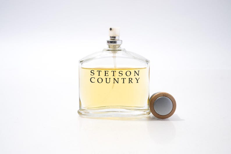 May include: A clear glass perfume bottle with a silver sprayer and a wooden cap. The bottle contains a light yellow liquid and is labeled "STETSON COUNTRY" in black text. The cap is next to the bottle.