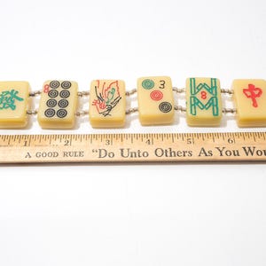 May include: A vintage bracelet featuring six rectangular Mahjong tiles. The tiles are cream-colored with black, red, and green symbols. The bracelet is approximately 7 inches long, as measured by a wooden ruler.