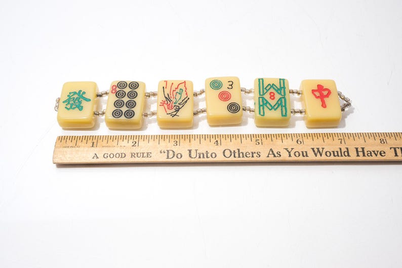 May include: A vintage bracelet featuring six rectangular Mahjong tiles. The tiles are cream-colored with black, red, and green symbols. The bracelet is approximately 7 inches long, as measured by a wooden ruler.