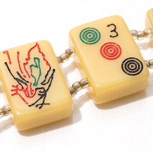 May include: Close-up of a bracelet featuring rectangular ivory-colored tiles, each adorned with colorful symbols and numbers. The tiles are connected by small, clear beads. The design includes red, green, and black details, with the number "3" visible.