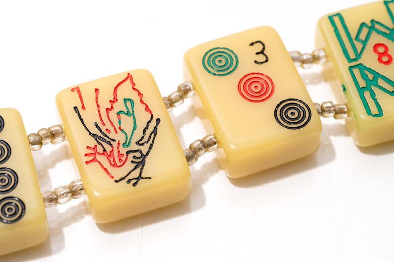 May include: Close-up of a bracelet featuring rectangular ivory-colored tiles, each adorned with colorful symbols and numbers. The tiles are connected by small, clear beads. The design includes red, green, and black details, with the number "3" visible.