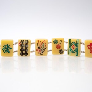 May include: A bracelet made of six rectangular, cream-colored Mahjong tiles. Each tile features a different design in red, green, and black. The tiles are connected by a string of small, clear beads. The bracelet is displayed on a white surface.