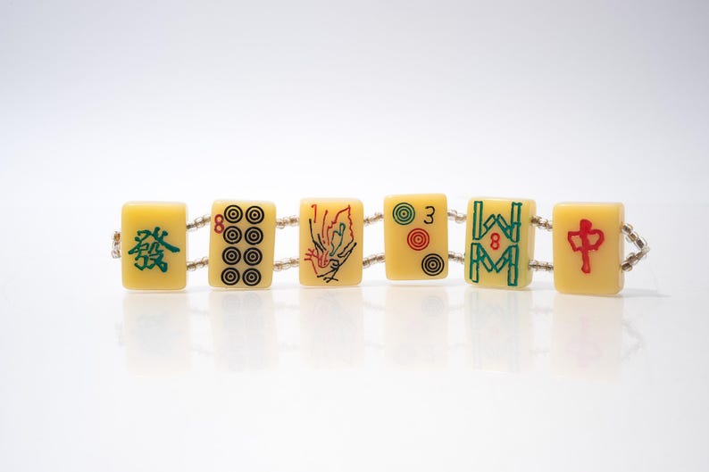 May include: A bracelet made of six rectangular, cream-colored Mahjong tiles. Each tile features a different design in red, green, and black. The tiles are connected by a string of small, clear beads. The bracelet is displayed on a white surface.