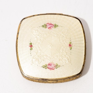 Puede incluir: Un estuche compacto cuadrado vintage con una superficie crema y un marco dorado. La superficie presenta un diseño floral en relieve con rosas rosas y hojas verdes. Un accesorio cosmético clásico.