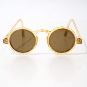 May include: Round sunglasses featuring a translucent, pale yellow plastic frame and temples. The lenses are a solid, medium-brown tint. The bridge is arched, connecting the two circular rims, and the temples are attached with small metal hinges.