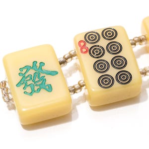 May include: A close-up of a bracelet featuring rectangular ivory-colored Mahjong tiles. The tiles are connected by small beads. Some tiles have black and red circular designs, while others feature teal Chinese characters.