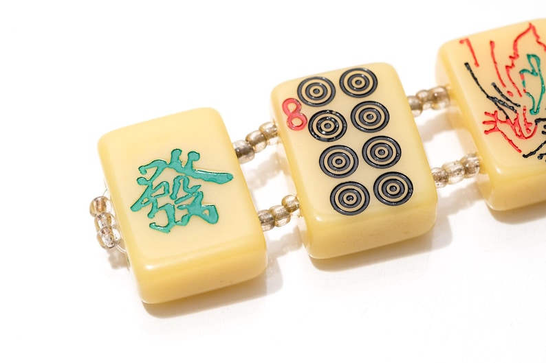 May include: A close-up of a bracelet featuring rectangular ivory-colored Mahjong tiles. The tiles are connected by small beads. Some tiles have black and red circular designs, while others feature teal Chinese characters.