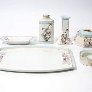 May include: A porcelain vanity set featuring a large rectangular tray, a small dish, a tall bottle with a stopper, a candlestick, a lidded jar, and a small bowl. Each piece is cream-colored with light blue trim, gold accents, and floral motifs.