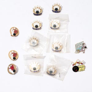 Pode incluir: Uma coleção de 11 pinos de boliche dourados. Os pinos têm vários designs, incluindo uma bola de boliche com um design de 200 club vermelho e branco, uma bola de boliche com um design de 200 club azul e branco e uma bola de boliche com um design de jogo limpo azul e branco. Os pinos estão em várias embalagens, incluindo sacos de plástico e caixas de papelão.