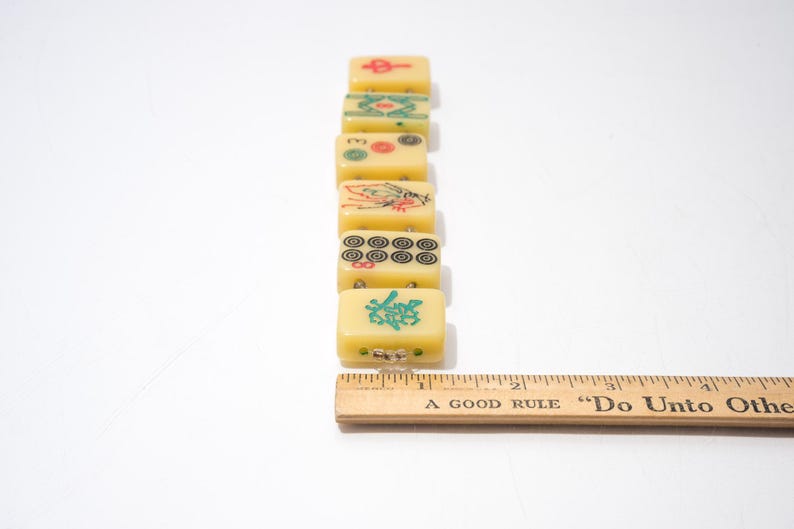 May include: A row of vintage yellow Mahjong tiles with various symbols in red, green, and black. A wooden ruler with the text "A Good Rule Do Unto Others" is next to the tiles. The tiles are rectangular and have a slightly translucent appearance.