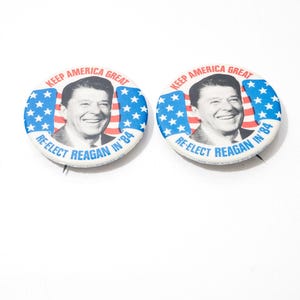 May include: Two vintage campaign badges. Each badge features a portrait of Ronald Reagan, with the text "KEEP AMERICA GREAT" and "RE-ELECT REAGAN IN '84". The badges have a blue and white star design.