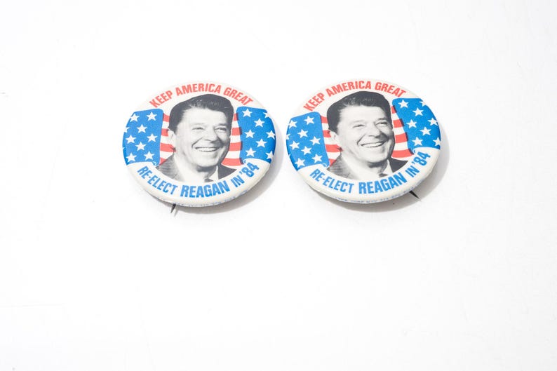 May include: Two vintage campaign badges. Each badge features a portrait of Ronald Reagan, with the text "KEEP AMERICA GREAT" and "RE-ELECT REAGAN IN '84". The badges have a blue and white star design.