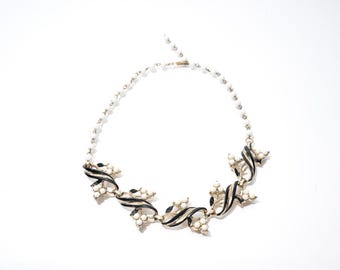 May include: A vintage necklace featuring a delicate design with black and white enamel accents. The necklace has a series of stylized floral elements connected by a strand of small, white beads. The metal has a gold tone, and the necklace is set against a white background.