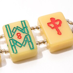 May include: Close-up of a bracelet featuring three ivory-colored Mahjong tiles. The tiles are connected by a string of small, clear beads. The tiles display red and teal symbols and numbers, including the number 8 and a Chinese character.