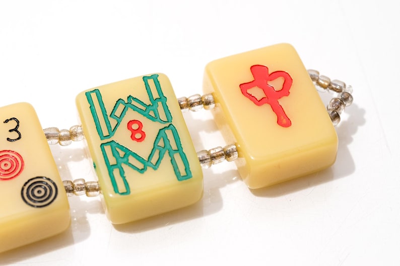 May include: Close-up of a bracelet featuring three ivory-colored Mahjong tiles. The tiles are connected by a string of small, clear beads. The tiles display red and teal symbols and numbers, including the number 8 and a Chinese character.
