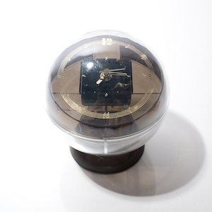 Puede incluir: Un reloj esférico transparente con una esfera cuadrada oscura y manecillas y números dorados. El reloj está encerrado en un globo transparente y se asienta sobre una base marrón oscuro. El diseño del reloj es retro y único.