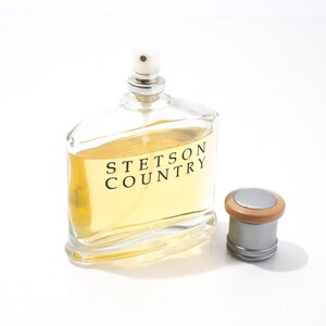 May include: A clear glass bottle of Stetson Country cologne with a silver cap and a wooden top. The bottle is filled with a light yellow liquid. The cap is next to the bottle.