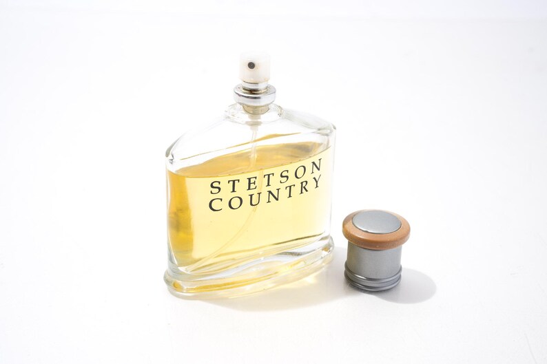May include: A clear glass bottle of Stetson Country cologne with a silver cap and a wooden top. The bottle is filled with a light yellow liquid. The cap is next to the bottle.