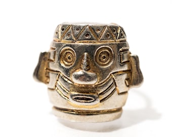 May include: A silver-toned metal ring with a stylized face design. The face has circular eyes, a small nose, and a smiling mouth. The ring has gold-colored accents and a geometric pattern on the top.
