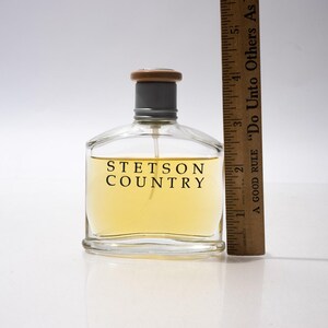 May include: A clear glass bottle of Stetson Country cologne with a light yellow liquid inside. The bottle has a gray cap and a wooden top. A wooden ruler is next to the bottle. The text "STETSON COUNTRY" is printed on the front.
