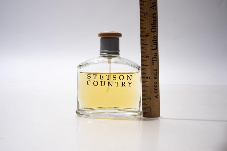 May include: A clear glass bottle of Stetson Country cologne with a light yellow liquid inside. The bottle has a gray cap and a wooden top. A wooden ruler is next to the bottle. The text "STETSON COUNTRY" is printed on the front.
