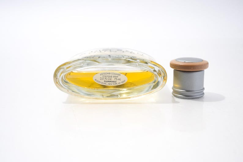 May include: An oval-shaped glass cologne bottle with a silver cap and a wooden ring. The bottle contains a yellow liquid and has a label that reads "Stetson Country Cologne Spray 2.5 FL OZ 75 ml".